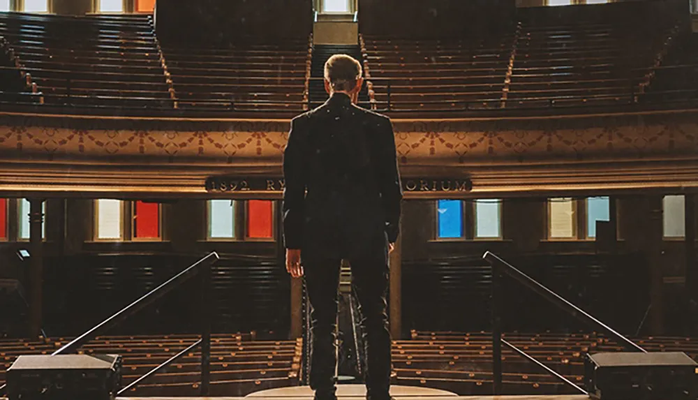 A person stands on a stage facing an empty auditorium with colorful stained glass windows