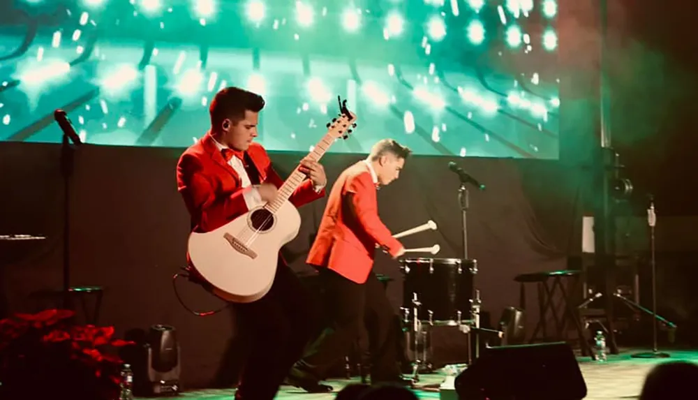 Two musicians in red jackets perform energetically on stage one playing an acoustic guitar and the other playing a drum against a vibrant illuminated backdrop