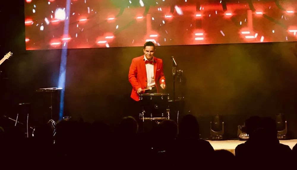 A drummer in a red jacket performs on stage with dynamic lighting in the background