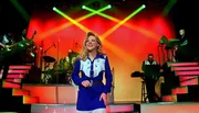 A performer in a blue and white outfit sings on a stage with a band in the background, illuminated by colorful stage lights.
