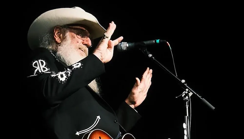 An older man with a beard wearing a cowboy hat and decorative jacket is performing on stage with a microphone and guitar