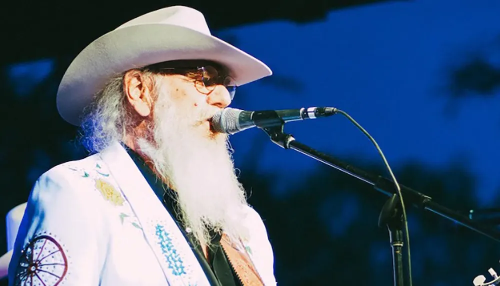 A man with a long white beard wearing a white cowboy hat and decorated jacket is singing or speaking into a microphone