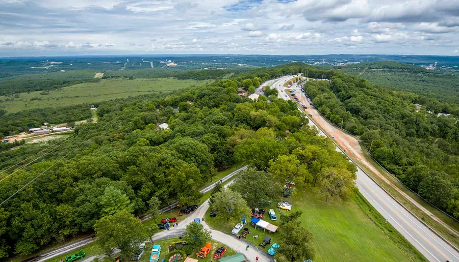 This image captures an aerial view of a curved highway slicing through a lush green landscape, with clusters of vehicles and infrastructure dotted alongside.