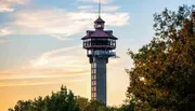 The image shows a tall observation tower with a distinctive hexagonal structure at the top, set against a backdrop of a sunset sky and surrounded by trees.