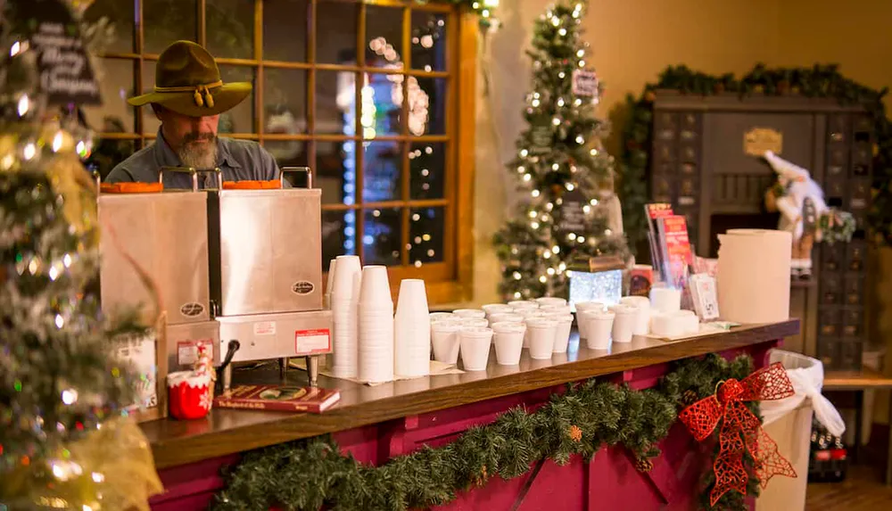 A person in a hat serves beverages from steel dispensers at a festive and decorated counter during a holiday event