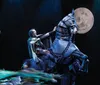 A person in theatrical attire rides a rearing horse puppet on stage set against a backdrop featuring a large full moon