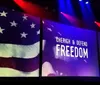 A person stands on stage beside a large screen displaying the American flag and the words Cherish  Defend Freedom