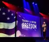 A person stands on stage beside a large screen displaying the American flag and the words Cherish  Defend Freedom