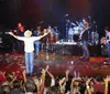 A musician in a cowboy hat stands with arms open on stage surrounded by a band performing for an enthusiastic crowd