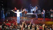 A musician in a cowboy hat stands with arms open on stage, surrounded by a band, performing for an enthusiastic crowd.