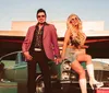 A man and a woman dressed in retro attire pose confidently in front of a vintage car with old-fashioned luggage nearby