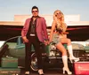 A man and a woman dressed in retro attire pose confidently in front of a vintage car with old-fashioned luggage nearby