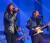 A woman is singing into a microphone while a man plays an electric guitar next to her on stage