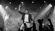 A band performs energetically on stage with a lively audience, as the lead singer raises his hand in excitement.
