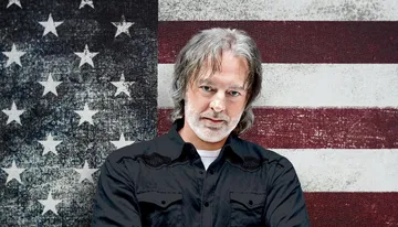 A man with gray hair and a beard stands in front of a weathered American flag.