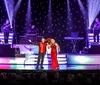A man and woman in formal attire are performing a dance on stage surrounded by Christmas trees and bright lights