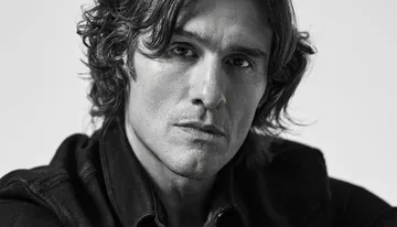 The image is a black and white close-up portrait of a person with wavy hair and a serious expression, wearing a dark jacket.