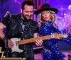 A man and a woman are energetically playing guitars on stage with a drum set and vibrant lighting surrounding them