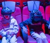 Several people are seated in red-lit chairs wearing virtual reality headsets appearing engaged and animated