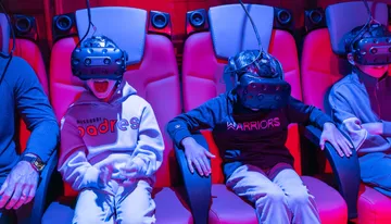 Several people are seated in red-lit chairs wearing virtual reality headsets, appearing engaged and animated.