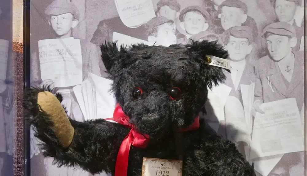A black teddy bear with a red ribbon and tag is displayed against a vintage photograph background