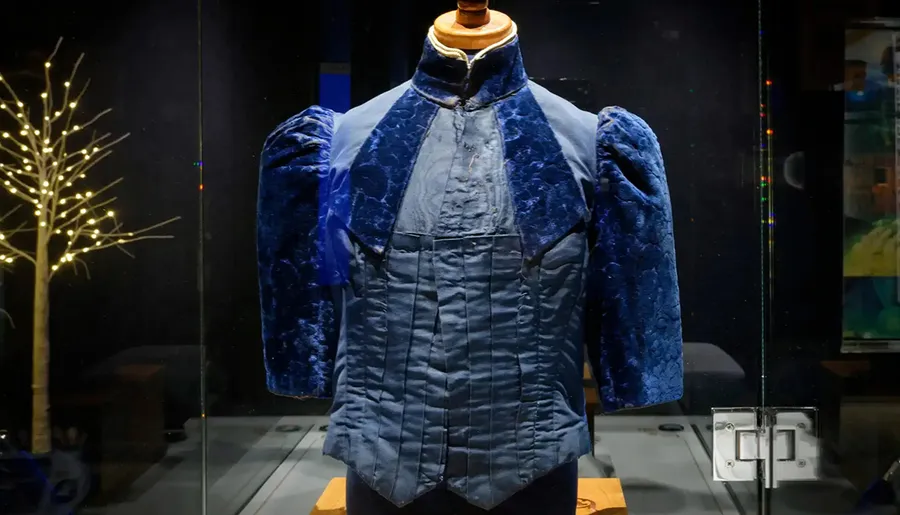 The image shows a blue, 19th-century men's jacket with a high collar and puffed shoulders displayed in a glass case, with a decorative tree light in the background.