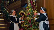 Two women in traditional maid outfits are decorating a lavishly adorned Christmas tree with ornaments near a grand staircase.