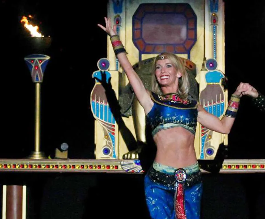 A person in Egyptian-themed costume is performing with a smile against a backdrop featuring Egyptian-style decorations and a lit torch.