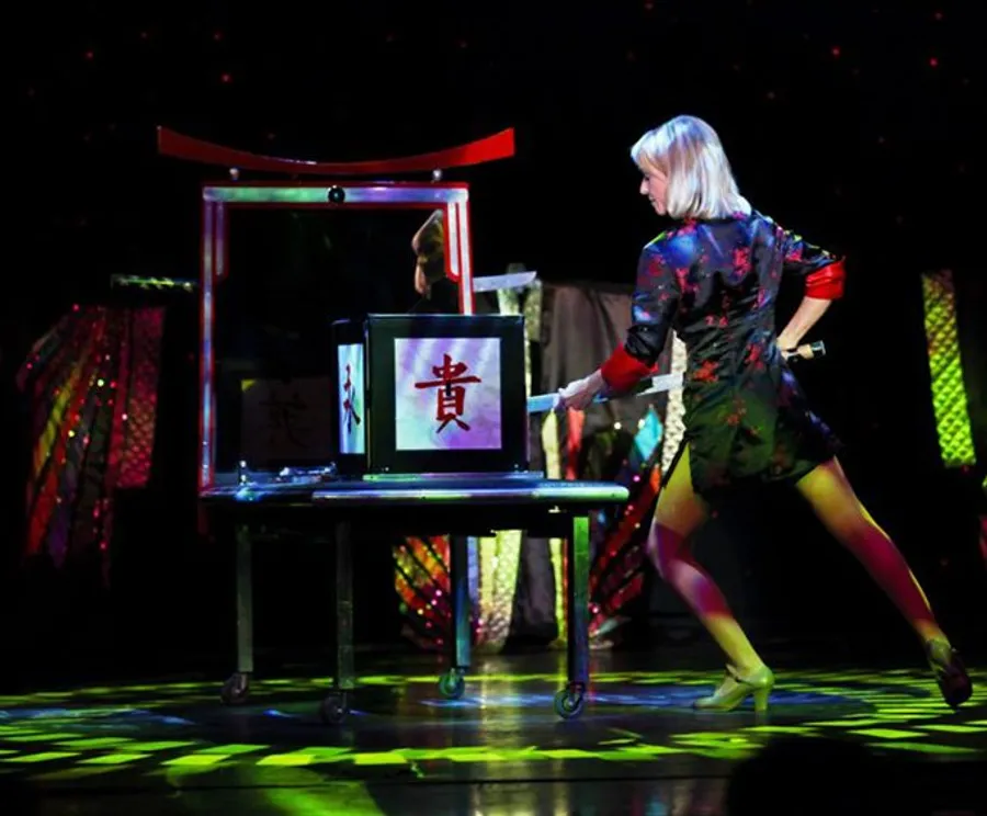 A performer is pushing a transparent box on a wheeled table with an Asian character displayed on it during what appears to be a magic show or theatrical performance.