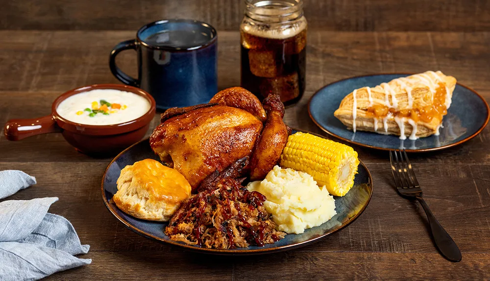 The image features a hearty meal with roasted chicken corn on the cob mashed potatoes biscuits pulled pork a bowl of soup a mug of beverage and a slice of pie all presented on a wooden table