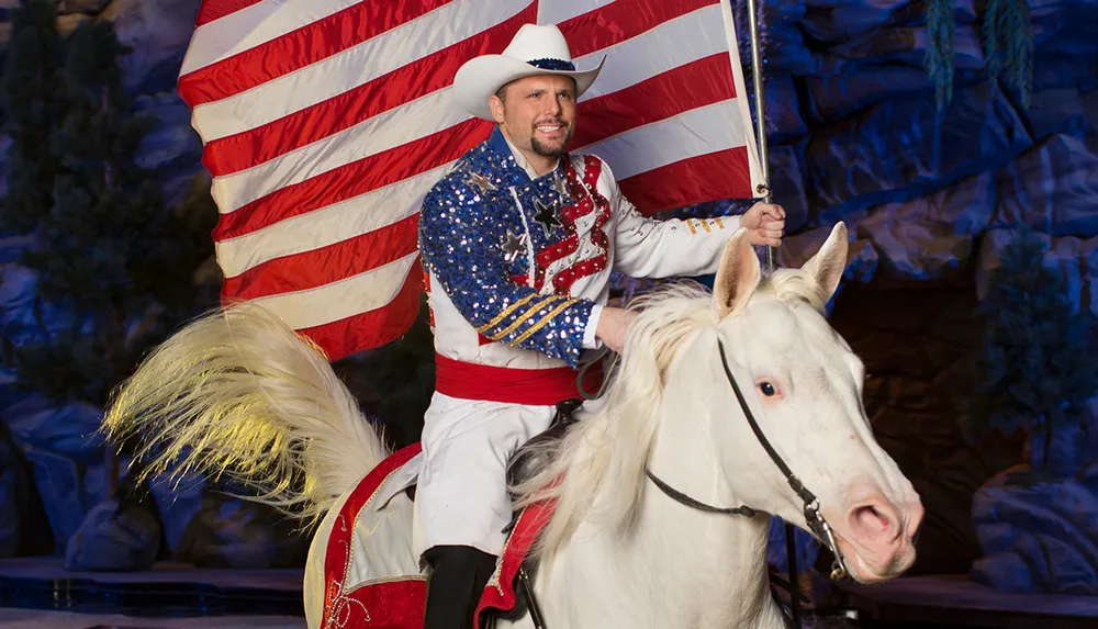 A man in a cowboy hat and a sequined patriotic outfit rides a white horse while holding an American flag