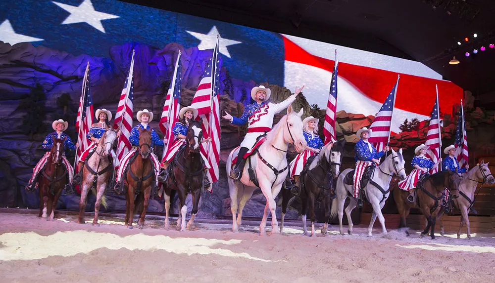 A group of people on horseback are dressed in patriotic outfits with American flags performing in front of a large backdrop featuring the stars and stripes