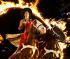 A performer rides two horses through a flaming ring in a dramatic lit-up setting