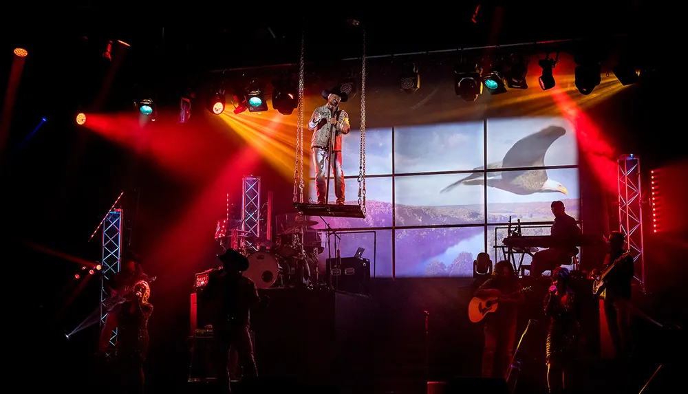 A musician performs on a raised platform among band members under colorful stage lights with an eagle displayed on the screen in the background