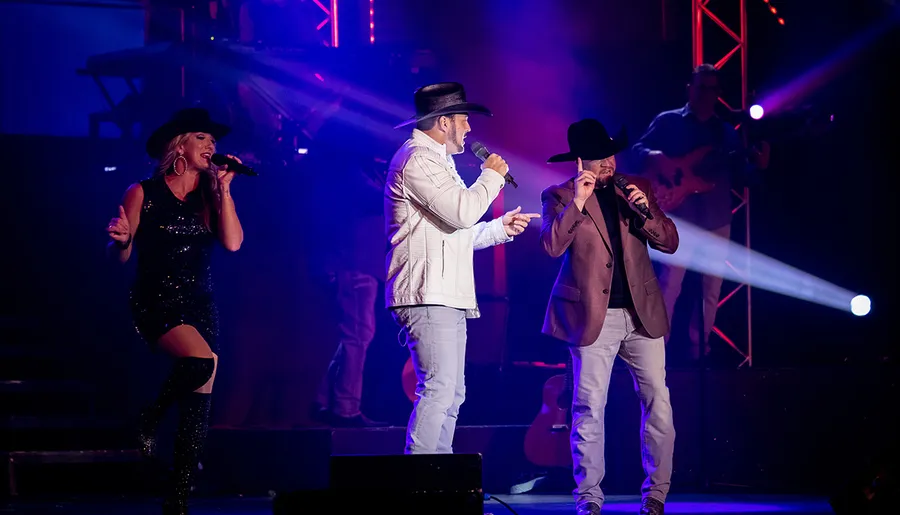 Two men and a woman in cowboy hats are performing on stage with colorful lights and a musician in the background.