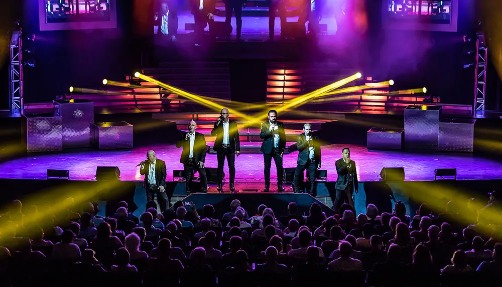 A group of performers are singing on stage in front of an audience with vibrant stage lighting enhancing the live entertainment experience