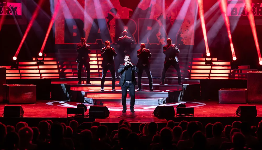 A singer is performing on stage with a microphone in hand accompanied by a group of backing vocalists under dramatic red stage lighting in front of an audience