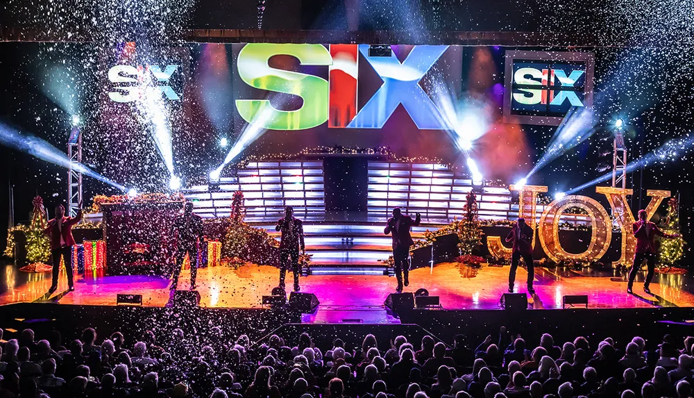 The image shows a vibrant live performance with performers on stage under colorful lights and confetti in front of an audience with large letters spelling SIX and JOY as part of the stage decor
