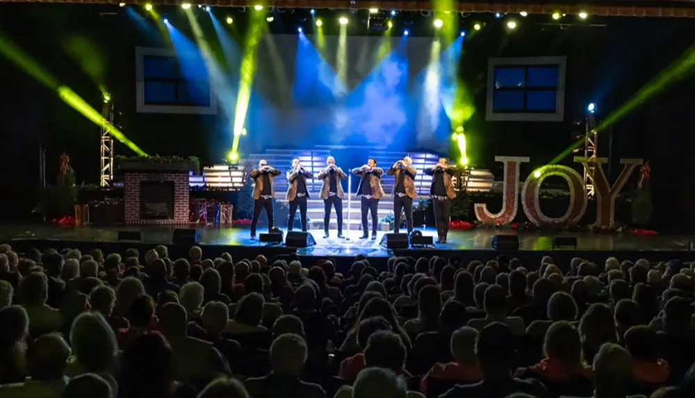 A group of performers is singing on stage in front of an audience with the word JOY prominently displayed at a concert or event