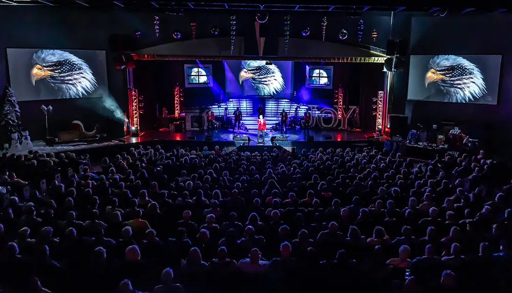 An audience watches a live performance on a stage adorned with impressive graphics of an eagle on screens with the event titled JOY