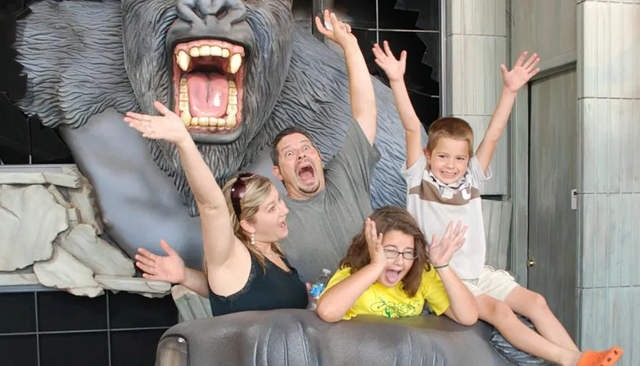 A family is playfully posing with their hands up as if scared, in front of a large, faux angry gorilla exhibit.