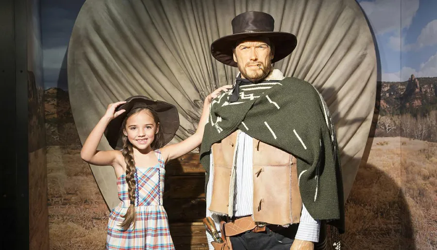 A young girl smiling at the camera poses with a lifelike figure dressed as a cowboy in front of a Western-themed backdrop.