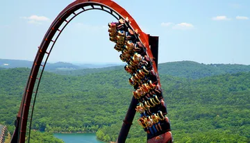 A roller coaster full of riders is ascending a steep loop with a lush green forest in the background.