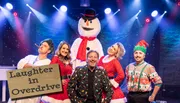 A group of people in festive attire pose with a snowman under stage lights, with a sign reading 