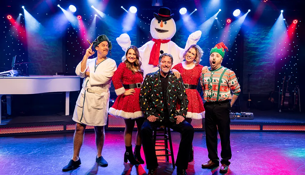 A group of people in festive holiday costumes including a snowman are joyfully posing on a stage with colorful lights