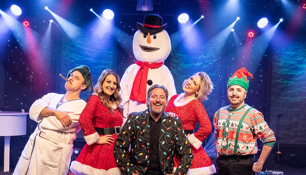 A group of people dressed in festive attire including a snowman pose happily on a stage with colorful lights