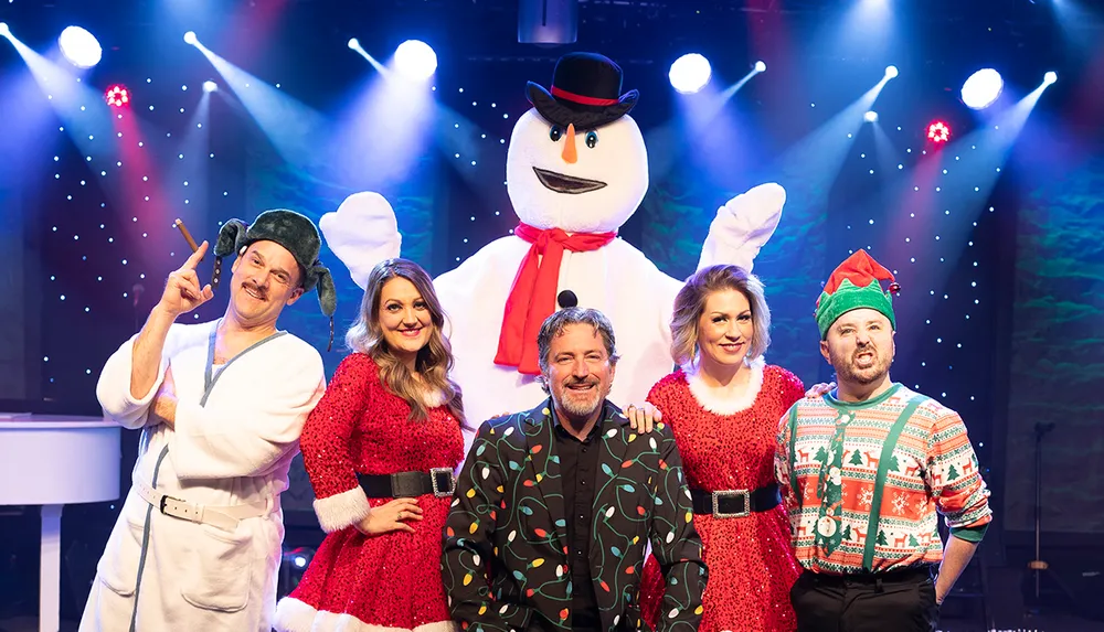 A group of people in festive holiday attire including a snowman costume are posing together on a stage with colorful lights in the background