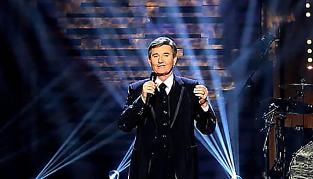 A person in a suit is performing on stage with a microphone highlighted by dramatic blue stage lighting and a backdrop that gives the appearance of a starry night