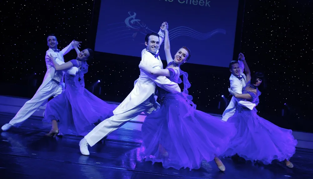 Dancers in white and purple attire elegantly perform under blue stage lighting with a starry backdrop and musical notes on a screen
