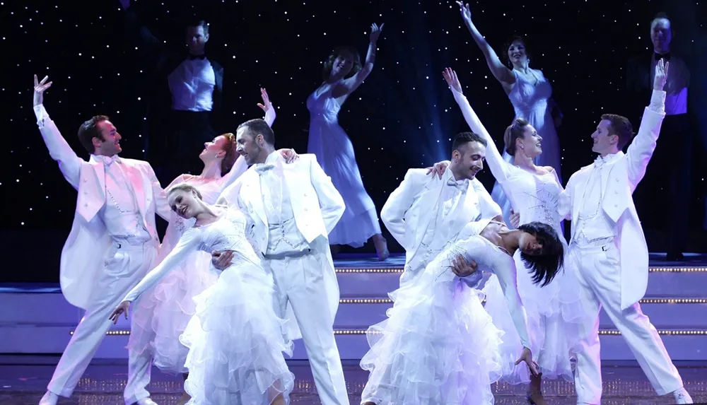Performers in white attire are elegantly dancing on a stage with a starry background creating a festive and graceful atmosphere